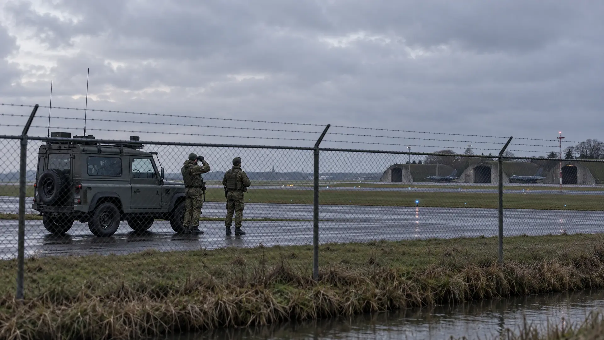 Two soldiers in camouflage uniform scanning an empty grey sky on a concrete apron beside a chain-link fence, with a patrol vehicle and the low hangars of a Dutch military airbase behind them